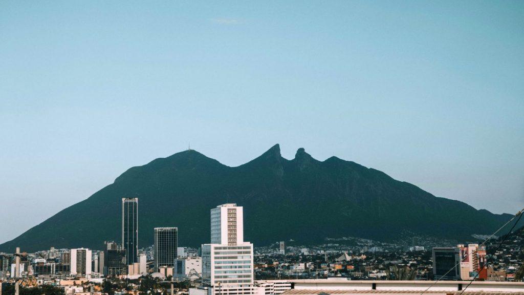 City skyline with tall buildings in front of a large green mountain range against a clear blue sky.