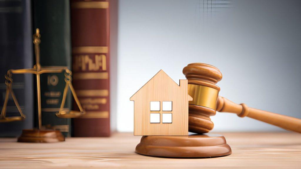 Wooden house model on a judge's gavel with scales of justice and law books in the background.