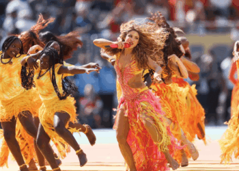 Performer in a pink fringed outfit sings into a microphone as teammates in yellow dresses dance on a stadium track.