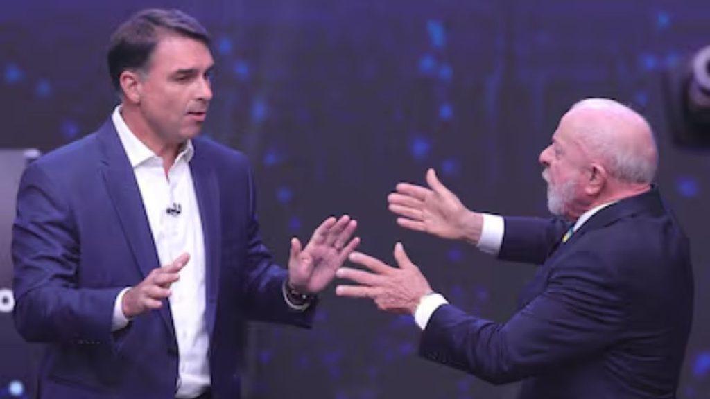 Two men in dark suits on a stage, facing each other with animated hand gestures during a discussion.] ,