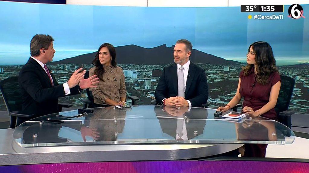 hechos-am-telediario-adrian-de-la-garza-alianza-2027jpg - HECHOS AM Four news anchors sit around a curved glass desk in a television studio, discussing a topic with a cityscape backdrop and a skyline behind them.