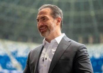 Man in a dark suit and white shirt smiling outdoors, with a blurred stadium background.